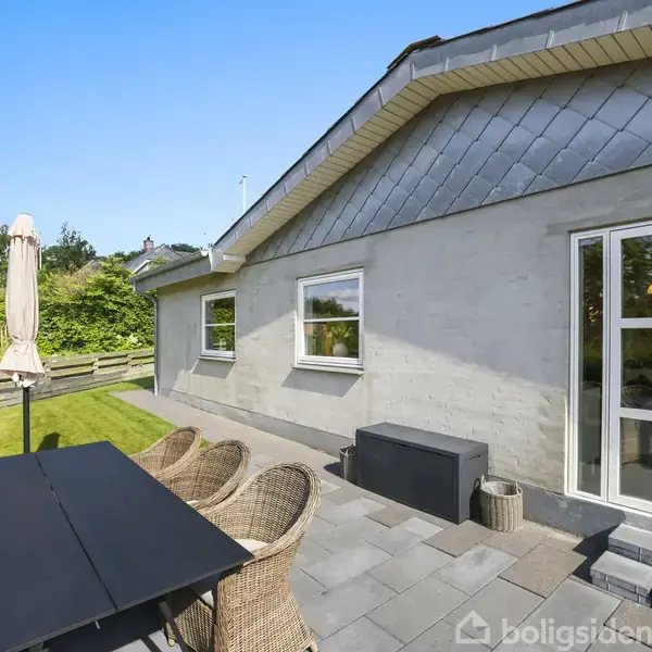 Et moderne hus med gråt tag ses fra terrassen, hvor et sort bord med flettede stole og en parasol står. Haven er grøn og omgivet af en lav træhegn.