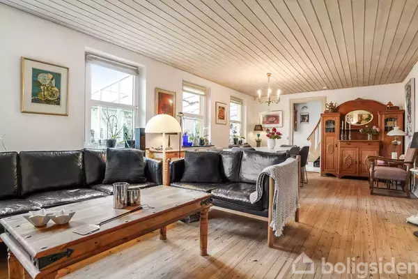 En stue med sort lædersofa og trækaffebord, placeret i et rum med trægulv og loft. Væggene er indrettet med billeder og kunst, og der er en skænk i baggrunden.