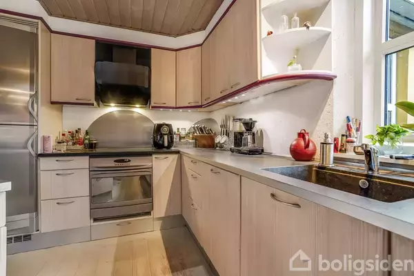 Køkken med lyse træskabe og stålfarvet ovn under et sort emhætte. På bordet står en kaffemaskine og en rød kedel. Vindue til højre, frisk plante ved vasken.