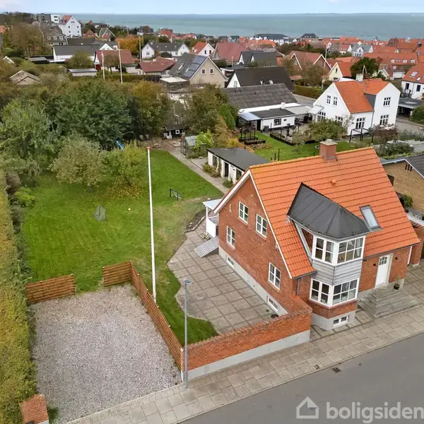 Murstenshuset står på en hjørnegrund med nyttehave og parkeringsplads. Omkringliggende er et boligkvarter med mange huse. I baggrunden ses havet.