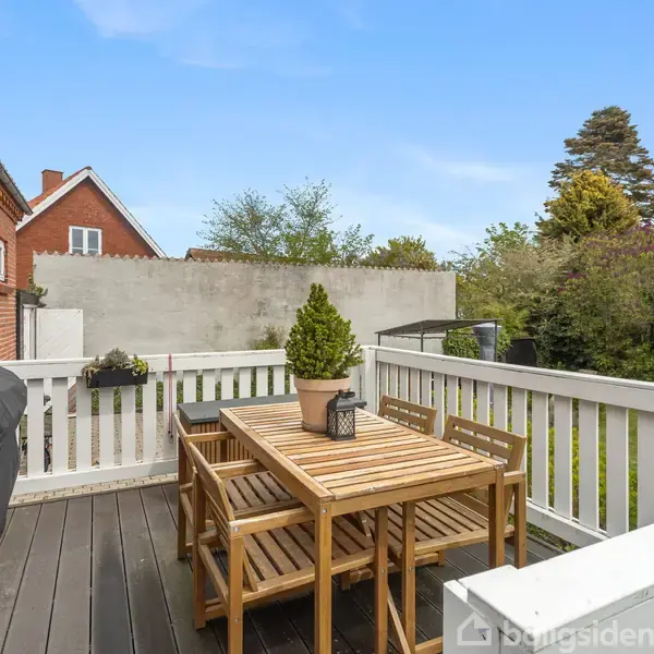 Træbord med seks stole står stille på en terrasse omkranset af hvidt hegn. En grill dækket af sort betræk står til venstre, omgivet af grønne planter og et murstenshus.