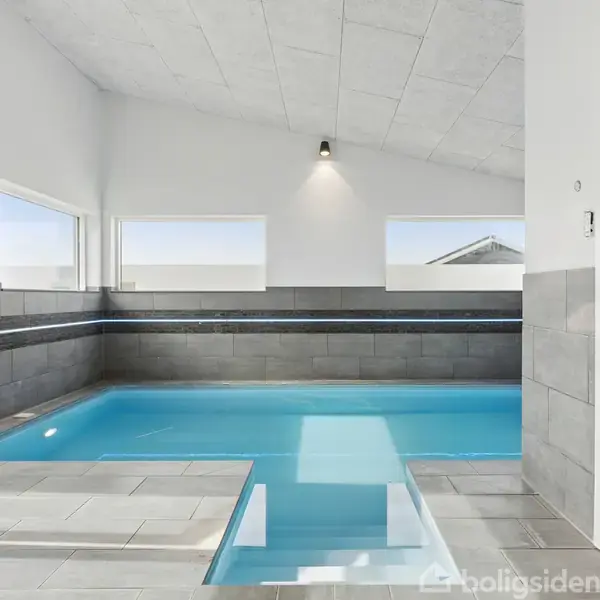 Indendørs swimmingpool med klart blåt vand omgivet af flisegulv og grå flisevægge. To store vinduer giver naturligt lys. Til højre er en dør til en sauna.
