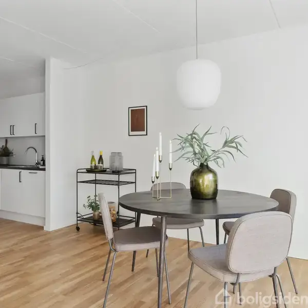 Rundt spisebord med fire stole står i en lys stue. Bordet er dekoreret med en grøn vase og lysestage. Ved siden af er et køkken med hvidt interiør og trægulve.