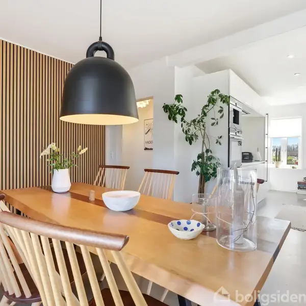 Langt træbord med stole omkring; hænger sort lampe over bordet. Væg med træpaneler i baggrund. Plante og vaser dekorerer bordet. Køkken kan ses i baggrunden.