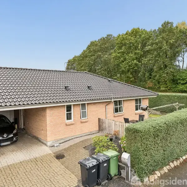 En murstensvilla med sort tegltag har en tilknyttet garage, hvor der holder en sort bil. Foran er en flisebelagt indkørsel med affaldscontainere, omgivet af grønne hække og træer i baggrunden.