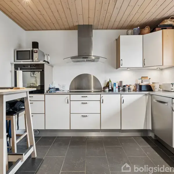 Et køkken med hvide skabe og en stål-emhætte. Bordpladen har forskellige køkkenredskaber. Et højt bord og stole står til venstre, og et vindue lukker lys ind.