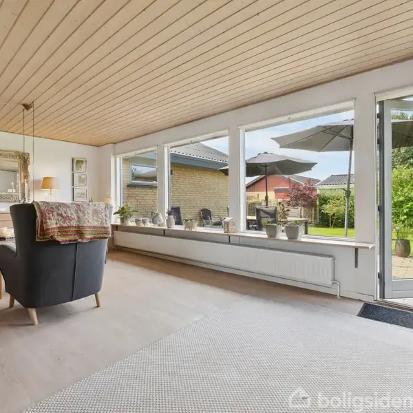 Stue med to lænestole vender mod store vinduer og en åben dør, der fører til en terrasse med parasoller og frodigt grønt haveområde.