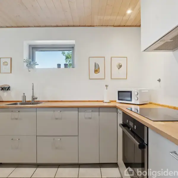 Køkken med lysegrå skabe, indbygget ovn og kogezone på træbordplader. Hvide fliser på gulv, hvidmalede vægge med indrammede billeder, og et lille vindue. Mikrobølgeovn og planter dekorerer bordet.