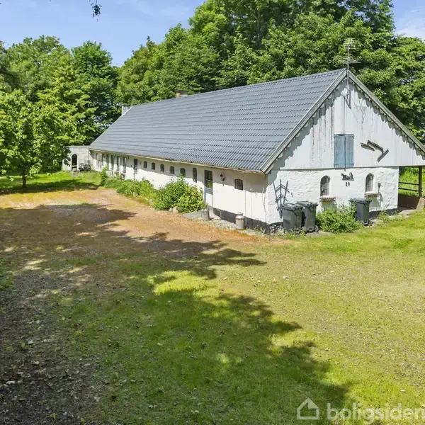 Hvid bygning med sort tag står i en skovklædt have. Omkring bygningen ses træer og grønne områder under en blå himmel.