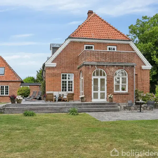 Et rødt murstenshus med terracottafarvet tegltag står i en have med græsplæne og flisebelagt terrasse. Haven er omkranset af grønne træer og buske.