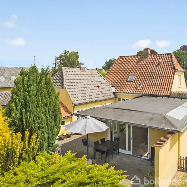 Et gult hus med rødt tegltag og overdækket terrasse med spisebord og parasol. Omgivet af velplejede haver og naboer i et villakvarter.