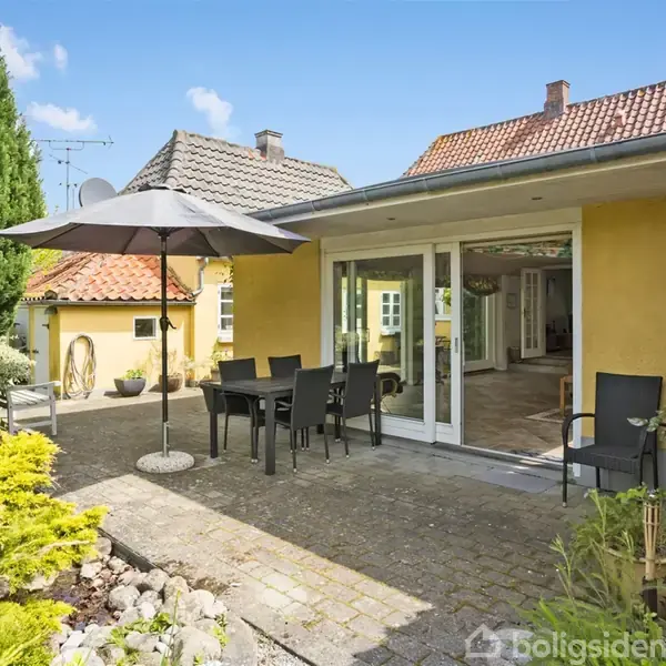 En terrasse med et mørkt bord og stole står under en parasol. Konteksten er en gulmalet bygning med vinduer og en have med grønne buske.