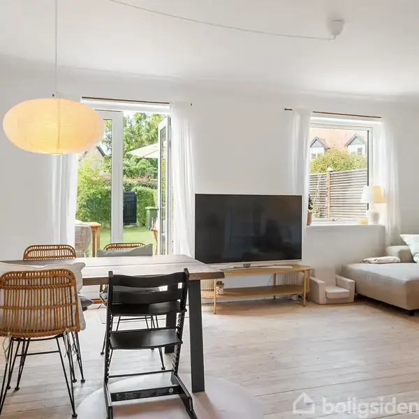 Stue med et spisebord og stole i træ, sofagruppe ved siden af et stort vindue, TV på et lavt bord, lysindstrømning fra haven gennem åbne døre.