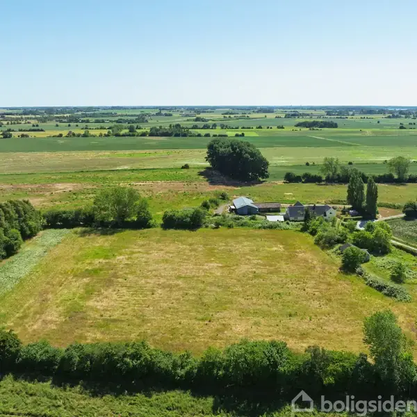 Græsmark ligger stille omgivet af landbrugslandskab. Et lille boligområde ses i baggrunden med grønne træer spredt omkring. Der er åbne marker og klar blå himmel.
