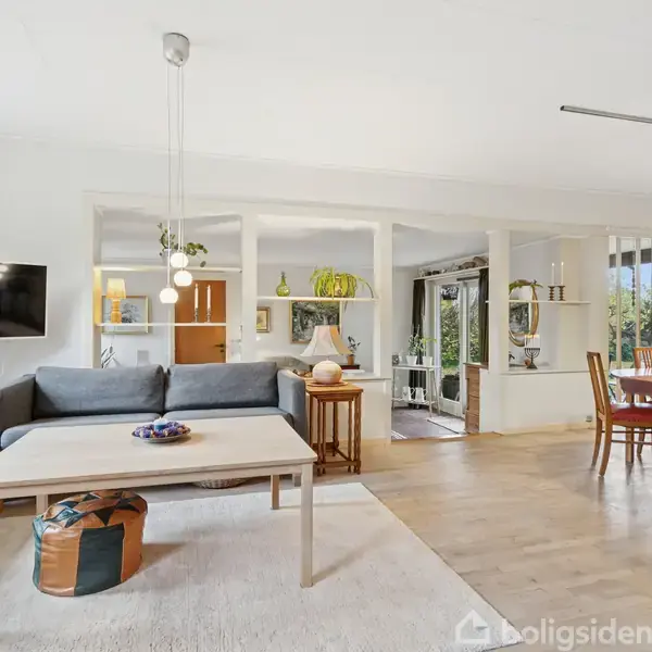 Stue med grå sofa, sofabord og tv til venstre. Spisebord med træstole til højre. Rummet er lyst med store vinduer og planter i baggrunden.