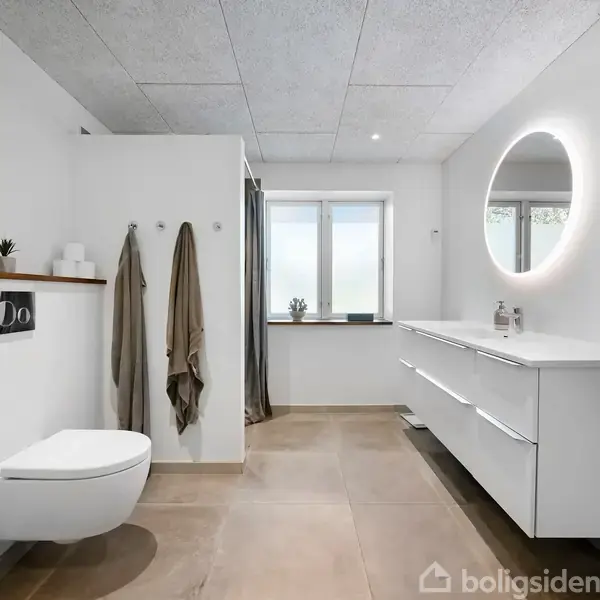 Toilet hænger på væggen; håndklæder hænger ved siden af. Badeværelse med hvidt interiør, stort spejl med cirkulært lys, skab og vindue med naturlig belysning.