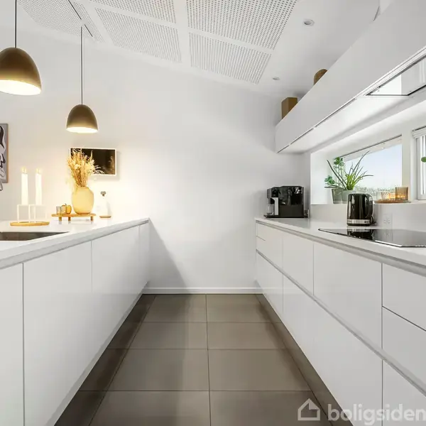 Et moderne køkken med hvide skabe og grå gulvfliser. To hængende lamper lyser rummet. På disken står planter, en kaffemaskine, og to lysestager. Vinduer til højre lader naturligt lys ind.