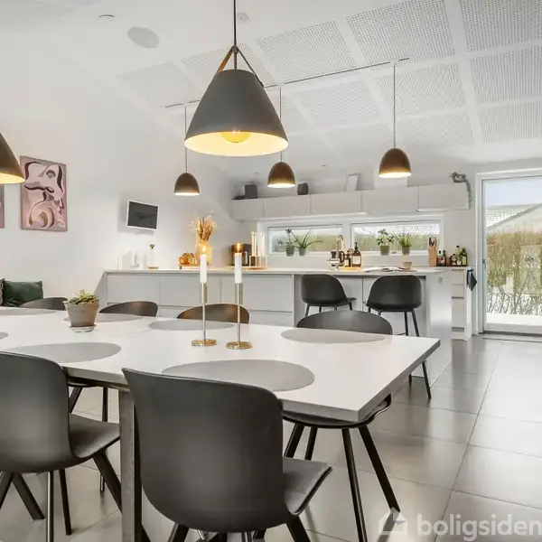 Spisebord med sorte stole står i et moderne køkkenalrum. Pendellamper hænger over bordet, og et stort vindue i baggrunden lader lys komme ind. Malerier hænger på væggen.