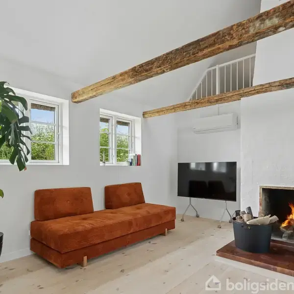 En stue med en brun sofa placeret ved siden af en stor grøn plante. Ild brænder i pejsen til højre, mens et TV hænger på væggen over en lys trægulv.