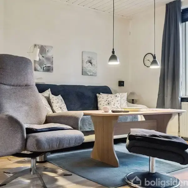 En grå lænestol med fodskammel står ved et sofabord i en stue. Sofaen bagved har puder. Rumoplysningen er hyggelig med to hængelamper og et vindue med gardiner.