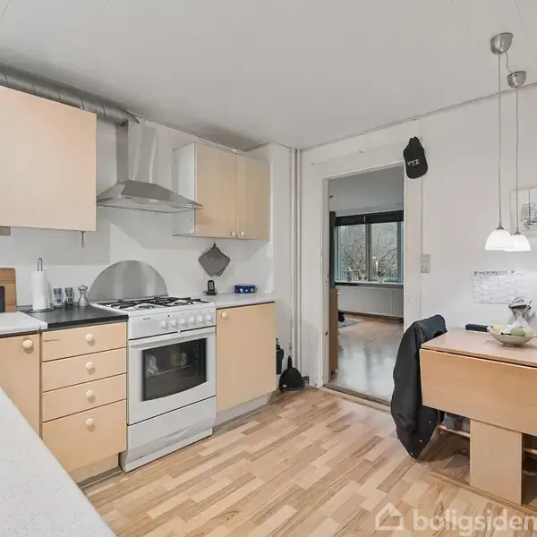 En køkken med lyse træskabe og komfur, hvor en emhætte er placeret over. Et spisebord i hjørnet ved siden af en lampe og stol. En dør åbner mod et tilstødende rum.