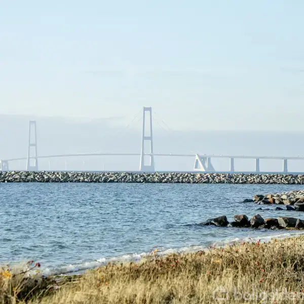 En lang bro strækker sig over vandet, med en stenet kystlinje i forgrunden og græsklædt område omkring. Klart vejr skaber en lys atmosfære.