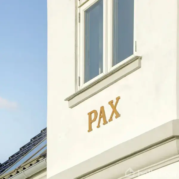 Bygning viser ordet "PAX" skrevet på facaden, nær et vindue. Området er solbeskinnet med en blå himmel i baggrunden og dele af nabotage synlige.