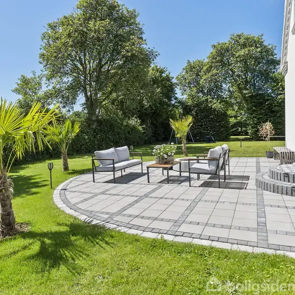 Terrasse med havemøbler står på flisebelægning foran et hvidt hus. Palmeplanter og grønne træer omgiver området, hvilket skaber en solrig og afslappende atmosfære i haven.