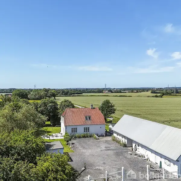 Hus med rødt tag står ved siden af en lang, hvid bygning. Omkring er der åbne marker og en vindmølle i baggrunden under en klar blå himmel.