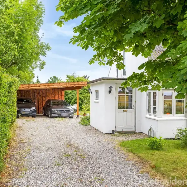 Et lille, hvidt hus med vinduer står ved siden af en grusvej. Til venstre er en carport med to parkerede biler. Omgivet af grønne træer og buske.