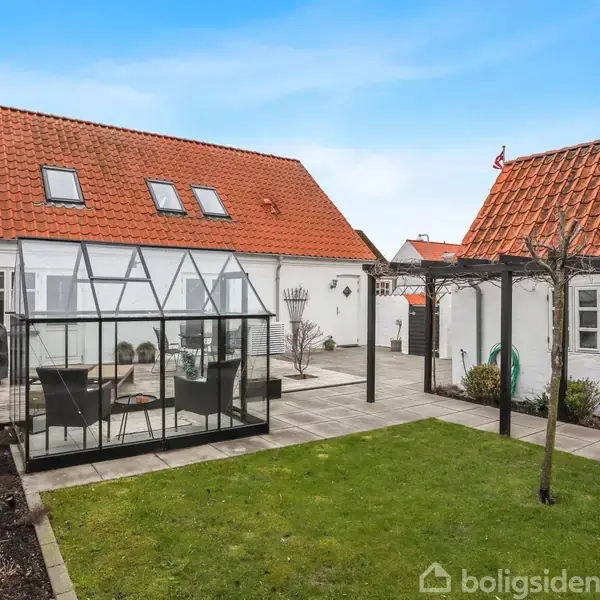 Drivhus med borde og stole står i en have omgivet af brostensbelægning. To hvidkalkede bygninger med røde teglstenstage omkranser gårdspladsen, i en hyggelig, velplejet setting.