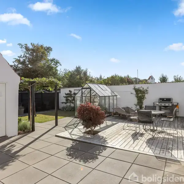 En gårdhave med træterrasse og havemøbler i et aflåst rum. Til venstre er der et hvidt skur. I midten står et lille drivhus omgivet af grønne planter.