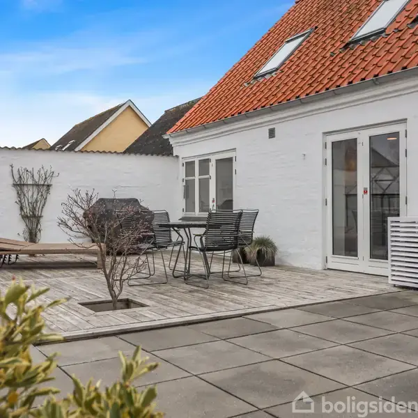 Hus med hvid facade og rødt tegltag har en terrasse med havemøbler, et glasdrivhus, og enkelte planter i krukker. Det er en solrig dag med blå himmel.