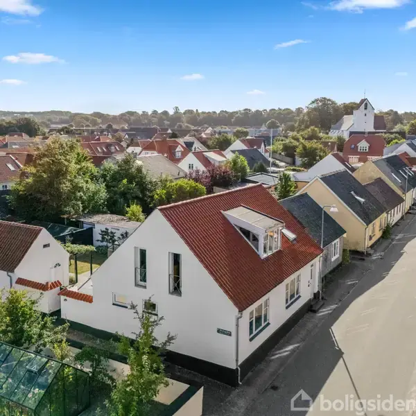 Hus med rødt tag ligger langs en stille gade omgivet af lignende huse og træer. Flaget foran huset viser national stolthed. Baggrunden viser en kirke og bakker.