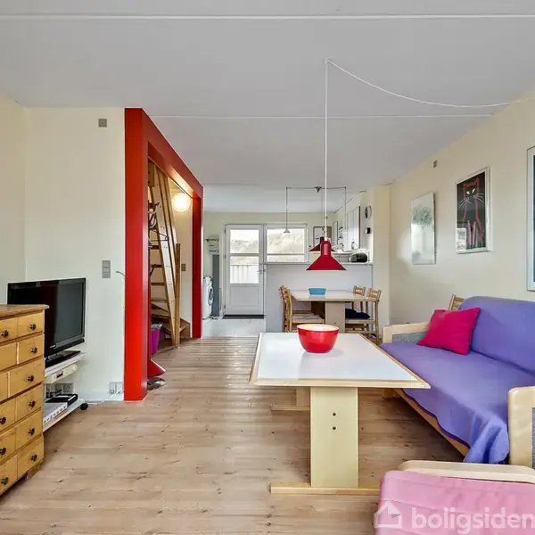 Stue med sofa dækket af blå og lilla tæppe, står ved et bord med rød skål. Fjernsyn på kommode til venstre. Lys hænger fra loftet over spisebord i baggrunden.