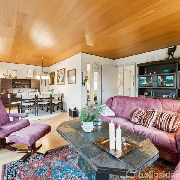 Stue med lilla sofa og lænestol står foran et sofabord med lys og planter. Spisestue i baggrunden. Træloft og store vinduer skaber en lys og indbydende atmosfære.