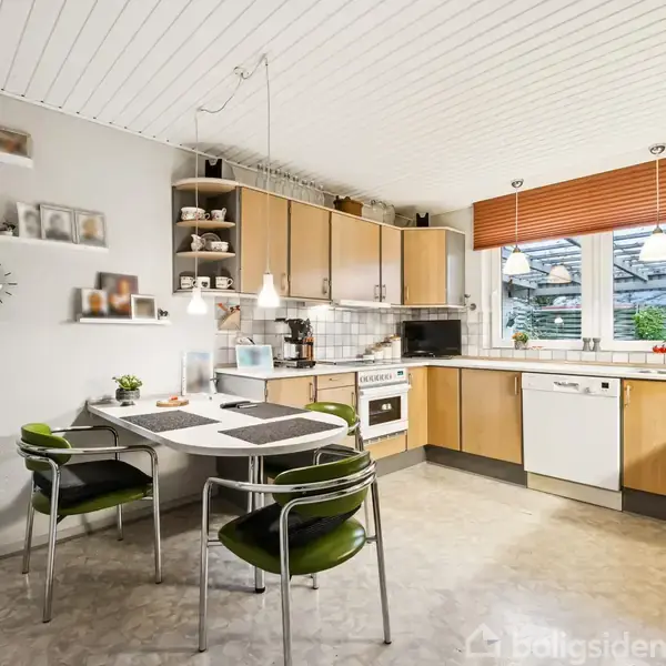 Et køkken med bord og fire stole, omgivet af træskabe og apparater som ovn og opvaskemaskine. Store vinduer med persienner giver udsigt til en have udenfor. Væggene har hylder og billeder.