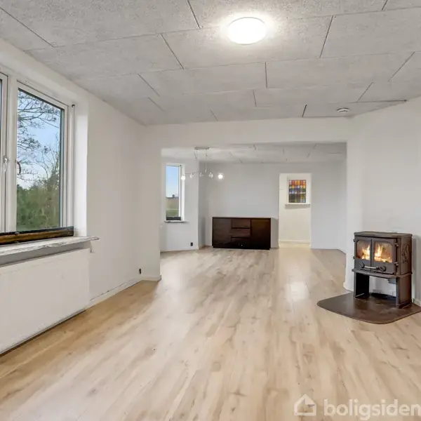 Et moderne køkken med sorte skabe og indbyggede ovne. Køkkenet har en kogeplade på en træbordplade. Rummet er lyst med lyse trægulve og store vinduer med udsigt til naturen.