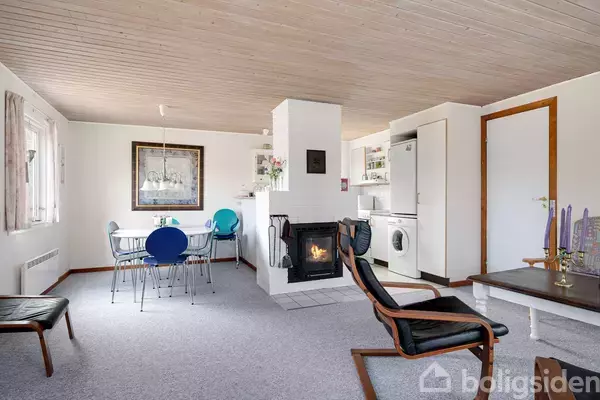 En hyggelig stue med en brændeovn, der brænder lystigt. I baggrunden er der et spisebord med stole. Vægge og loft fremstår i lyse trætoner, hvilket skaber en varm atmosfære.