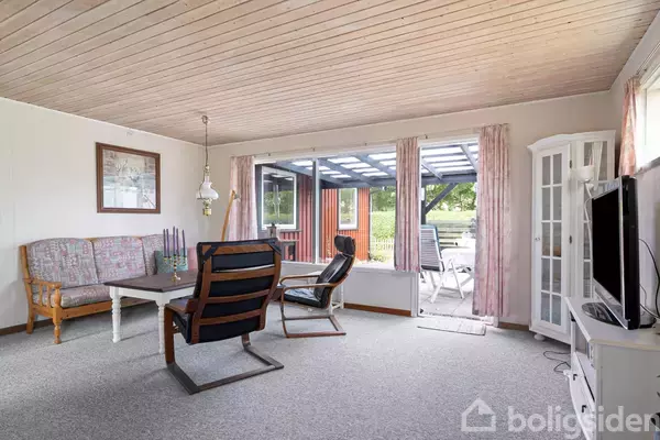 En stue med sofa og lænestole omkring et sofabord. En fladskærms-tv står på en skænk. Store vinduer og døre åbner ud til en overdækket terrasse med havemøbler.