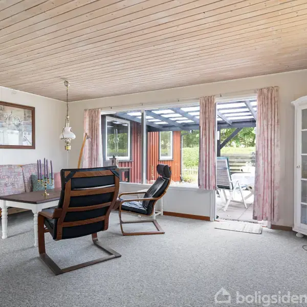 En stue med to stole og en sofa omkring et bord. Store vinduer viser en overdækket terrasse udenfor. Væggene er lyse, og der står en høj, hvid reol ved hjørnet.