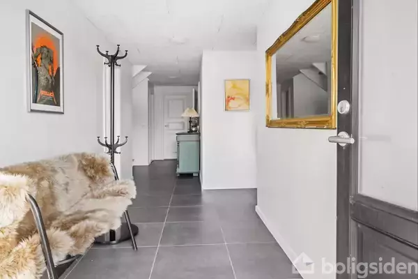 En gang med en pelsdækket bænk står langs væggen. En knagerække ved siden af bænken. På væggen hænger en stor, gylden ramme samt flere kunstbilleder.