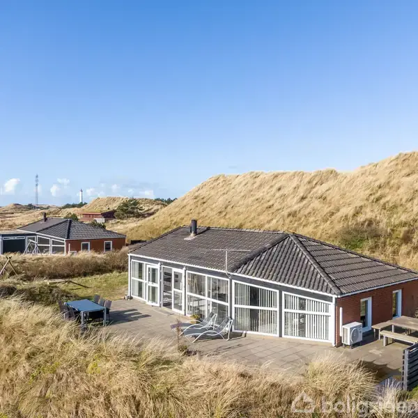 Et moderne hus står stille omgivet af klitter og græs. Store vinduespartier vender mod en solrig terrasse. I baggrunden ses flere huse og en mast mod en klar, blå himmel.