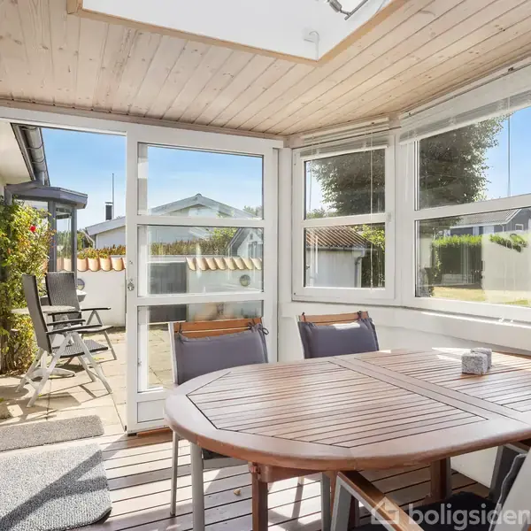 Et bord med stole står i en lys, indendørs veranda med store vinduer. Udenfor ses en terrasse med havemøbler og en udsigt til en have samt nabohuse.