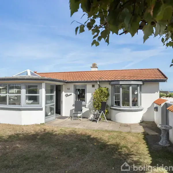 Huset har et fladt tag med orange tegl, store vinduer og en indgang. Det stå på en grøn græsplæne med træer i baghaven og en havekrukke ved siden af døren.