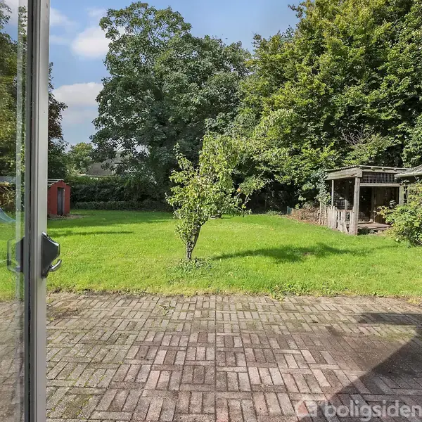 En glasdør åbner ud mod en flisebelagt terrasse og en græsplæne med træer og buske. Til venstre er et rødt skur, til højre en overdækket struktur omgivet af grønt.