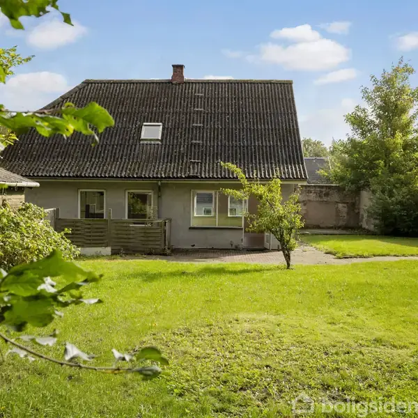 Hus med mørkt tag står i en have med grøn græsplæne og enkelte træer. Omgivet af andre bygninger og vegetation under en skyfri himmel.