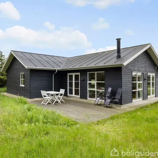 Et sort træhus med firehjørnet tag står på en træterrasse. To sorte liggestole og et lille hvidt bord med stole er placeret udenfor. Græsplæne omgiver huset.