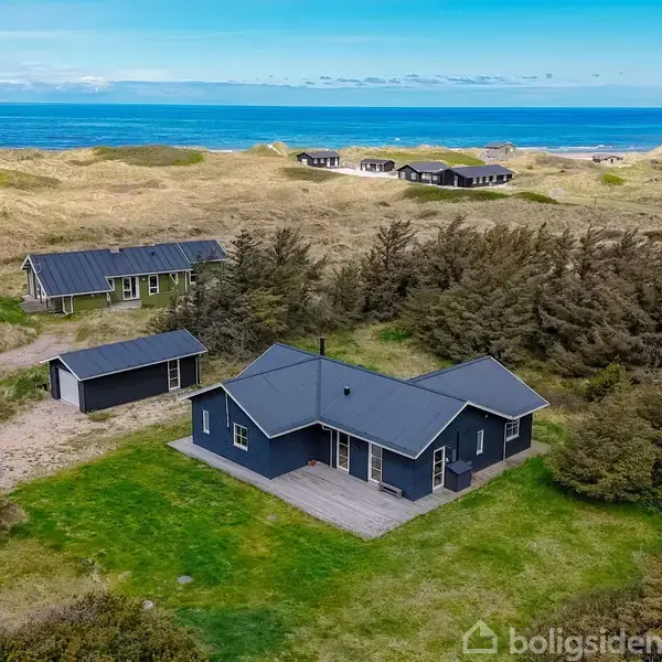 Et mørkeblåt hus står stille på en grøn græsplæne omkranset af buske. I baggrunden ses klitter og havet under en klar blå himmel.