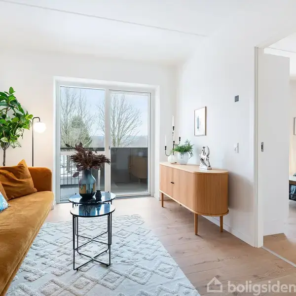 En gul sofa står ved siden af en grøn plante i en stue med en glasdør, der fører til en altan. En kommode står til højre, med et soveværelse synligt bagved.